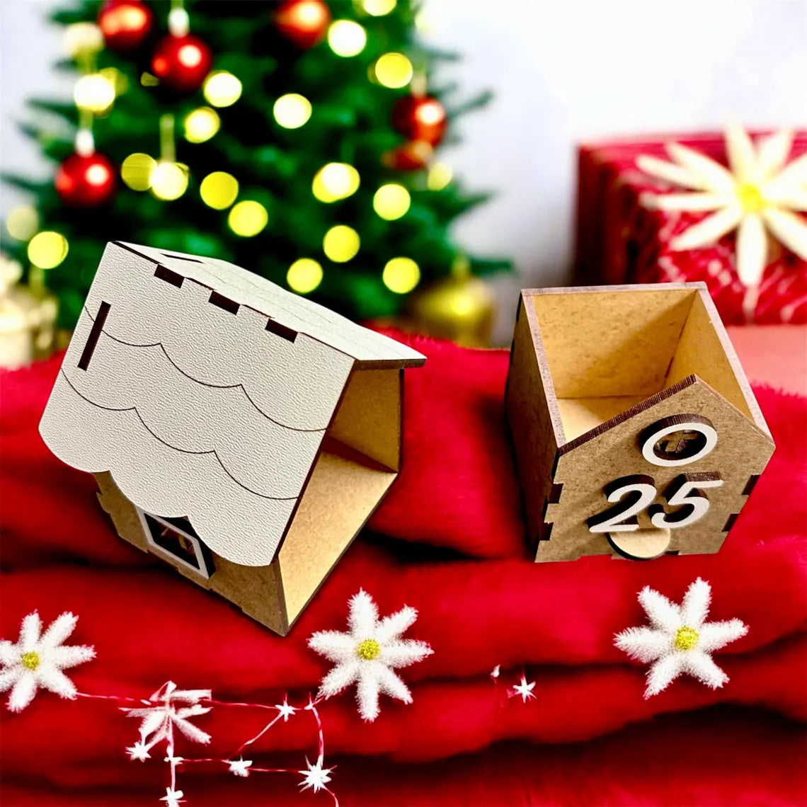 Two small cardboard houses on a red surface with a Christmas tree and gift in the background.