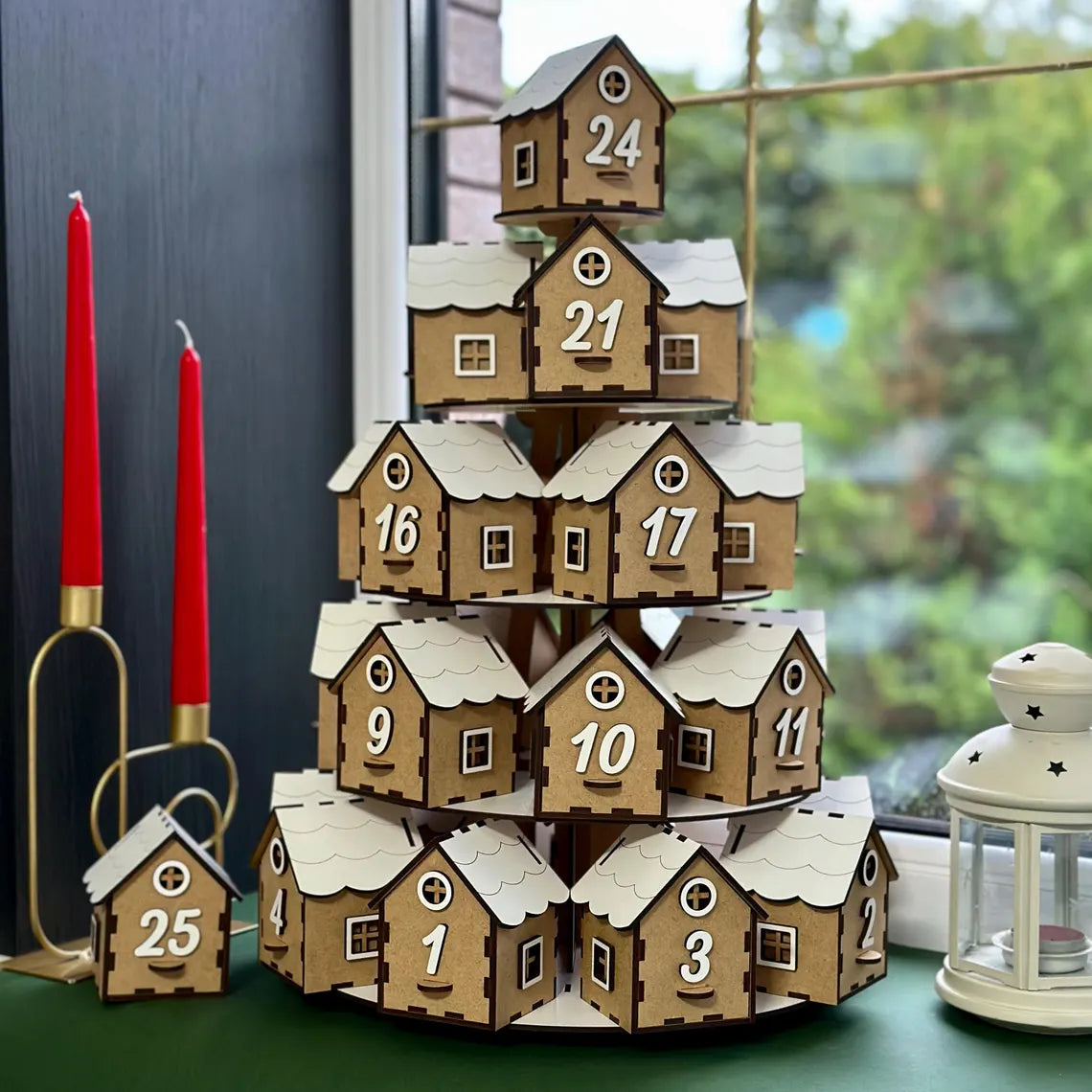 Wooden advent calendar with house designs and numbers, placed on a surface with candles and a lantern.