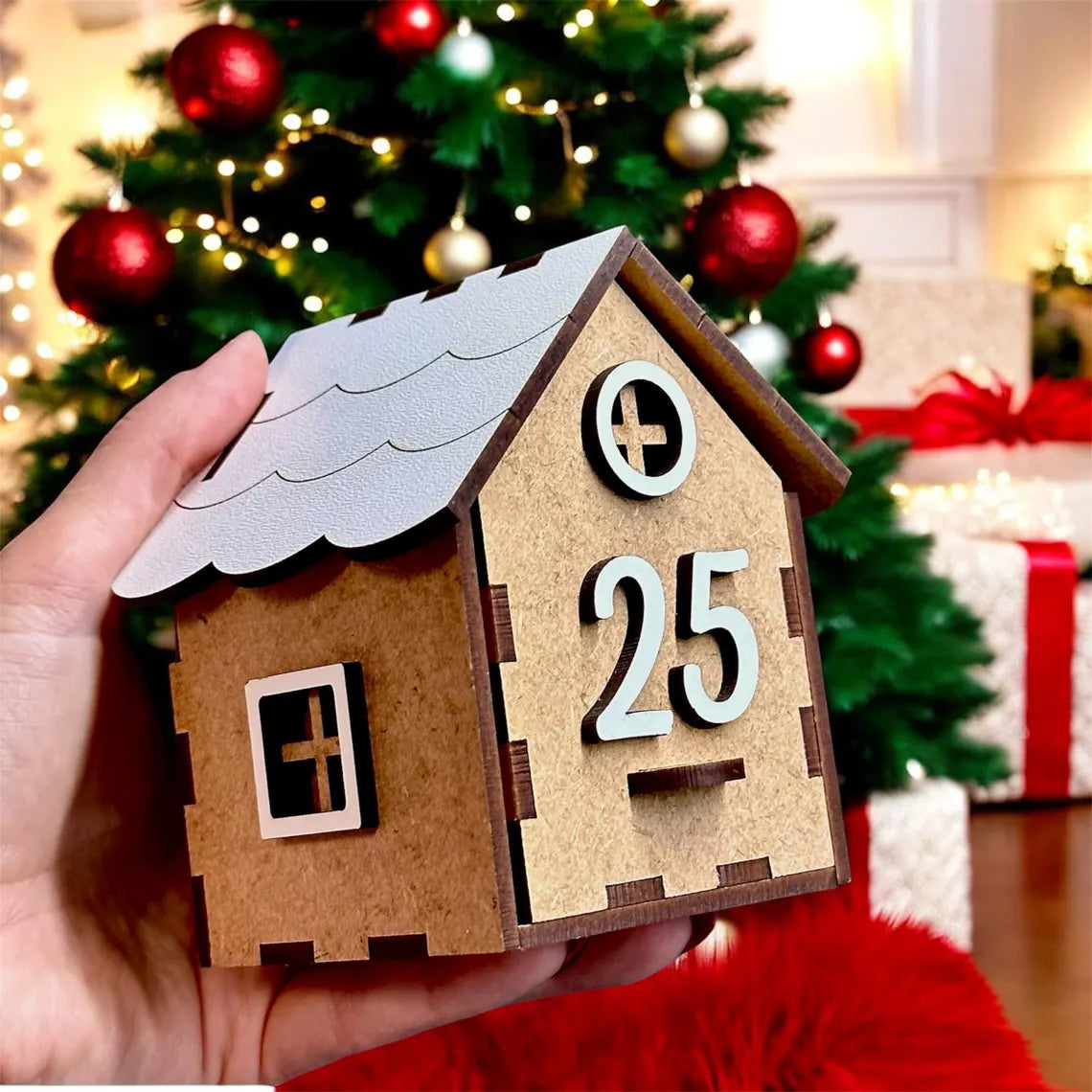 Wooden house-shaped Advent calendar with number 25, held in front of a Christmas tree and presents.