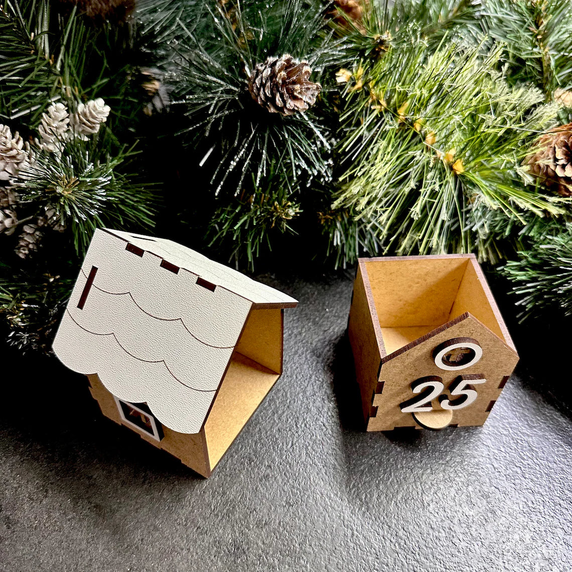 Two wooden house-shaped boxes with '25' on a gray surface with a Christmas tree in the background.