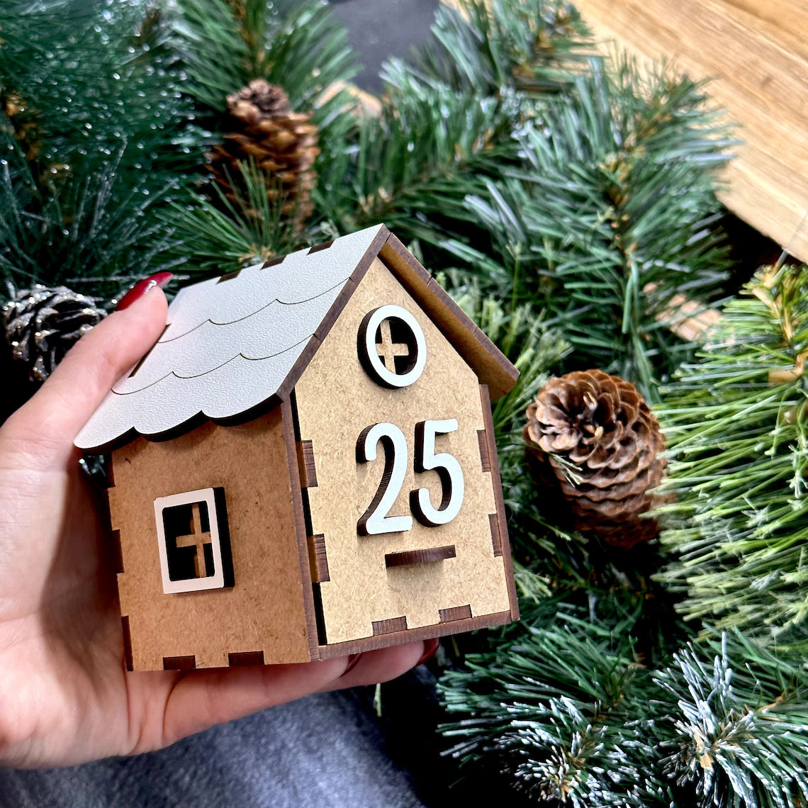 Small wooden house ornament with the number 25, held in front of Christmas tree branches.