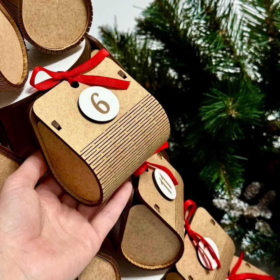 Wooden advent calendar with red ribbons and numbers, held by a hand with a Christmas tree in the background.