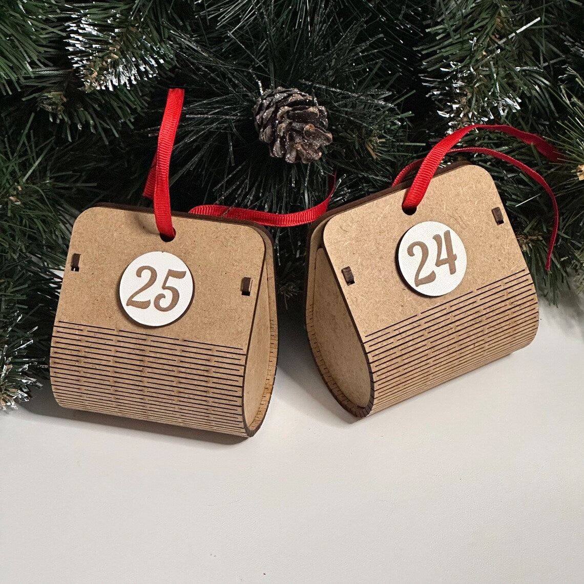 Two cardboard advent calendar doors with numbers 25 and 24 hanging from red ribbons against a Christmas tree background.