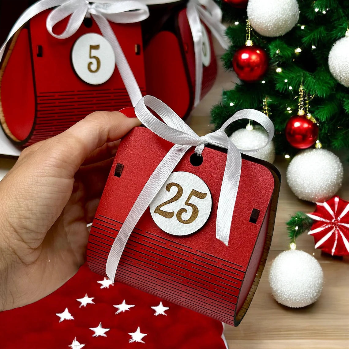 Red advent calendar with white ribbon and number 25, held by a hand, Christmas tree and decorations in the background.
