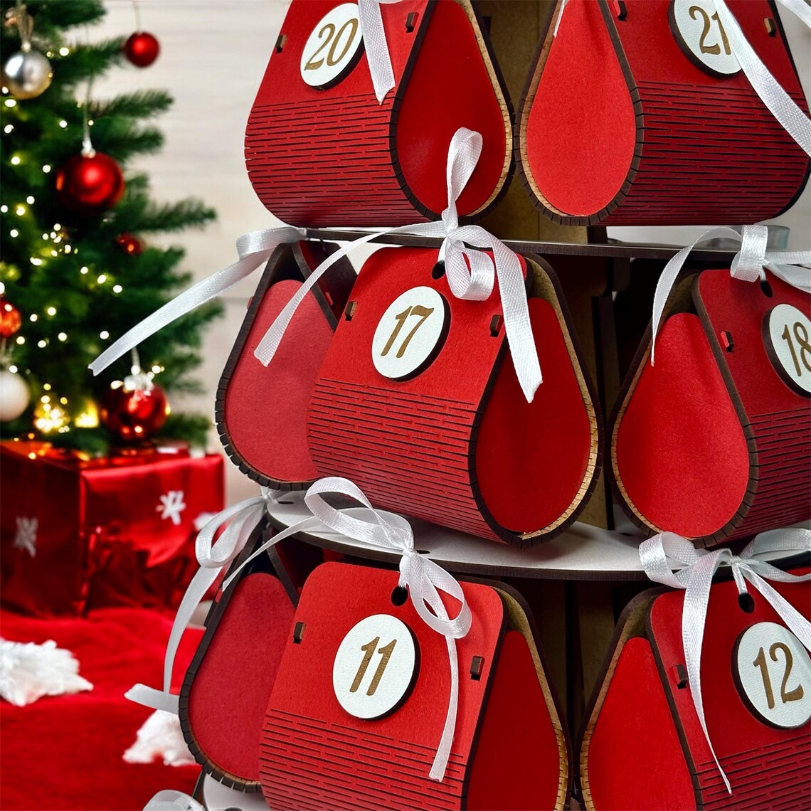 Red wooden advent calendar with numbered boxes in a festive setting with Christmas tree and presents.
