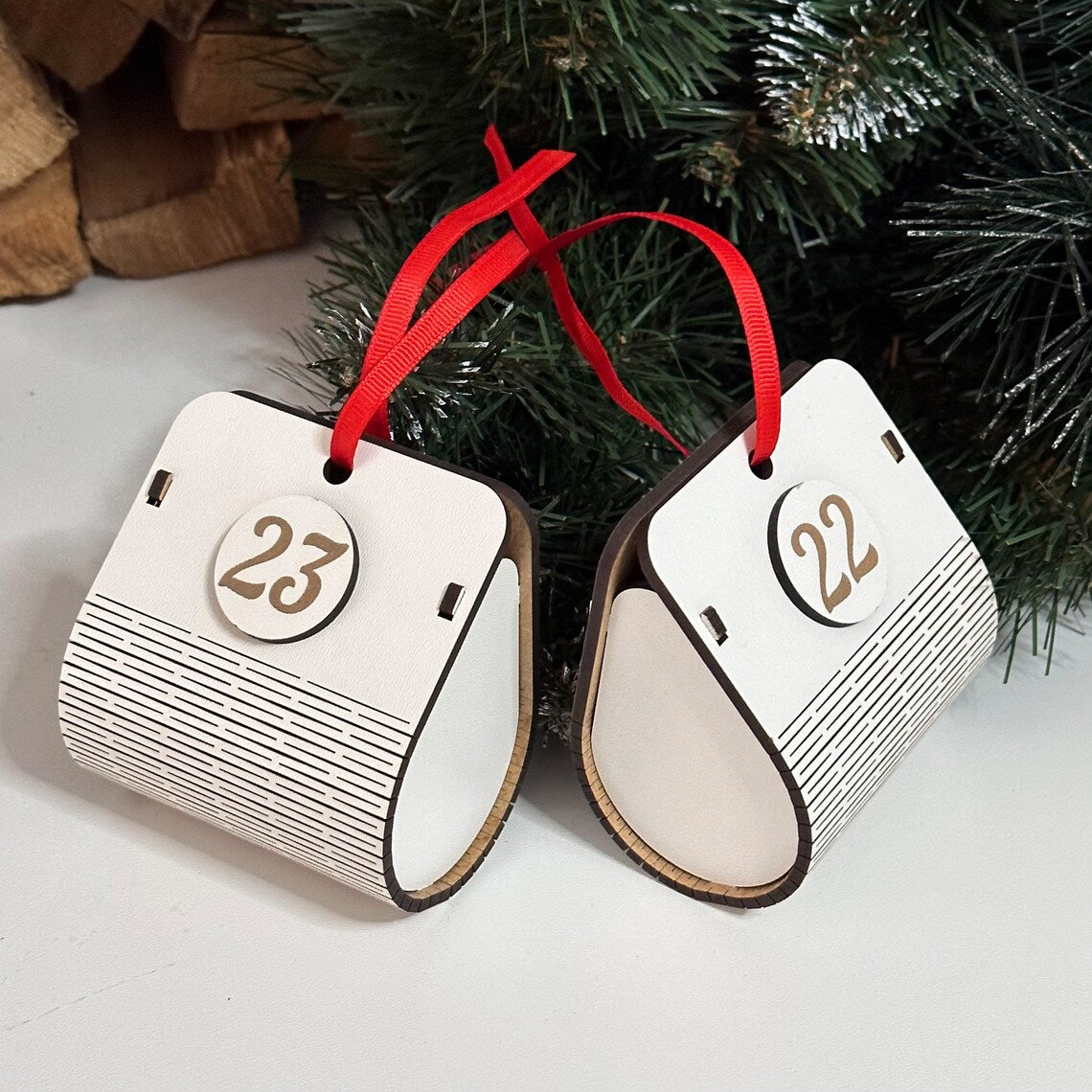 Two wooden advent calendar pieces with numbers 22 and 23, hanging from red ribbons against a Christmas tree background.