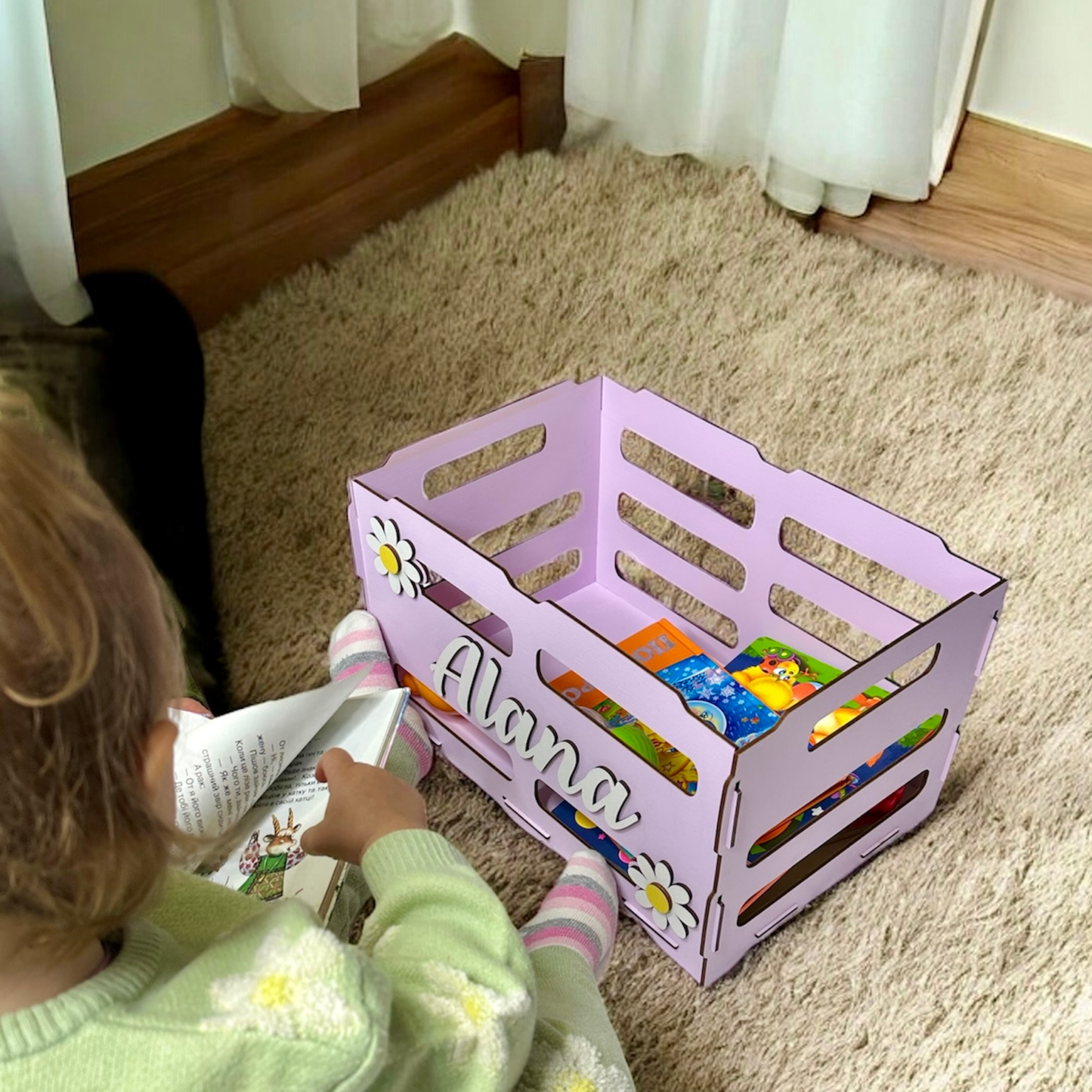 Child reading a book next to a pink storage crate with 'Alana' on it, on a carpeted floor.