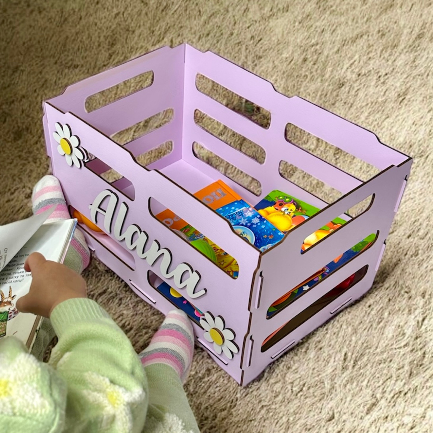 Child reading a book next to a purple storage crate with 'Alana' on it, filled with colorful books.
