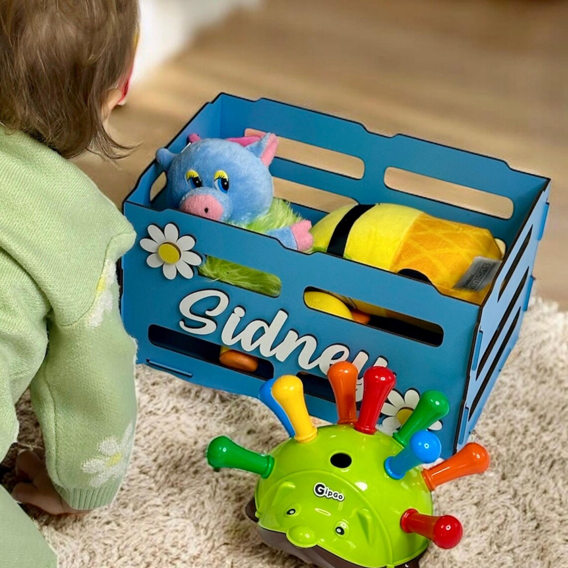 Child playing with toys inside a blue toy chest labeled 'Sidney' on a carpeted floor.