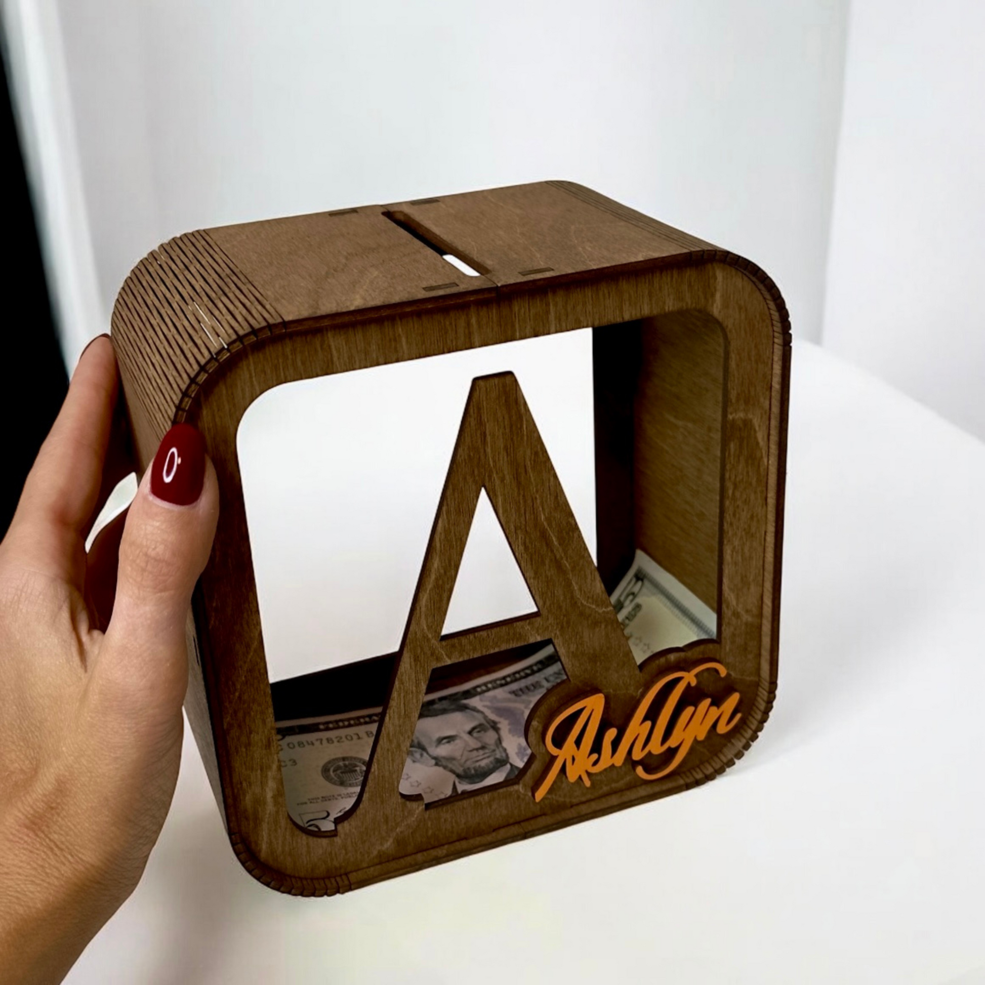 Wooden money box with 'Ashlyn' engraving, filled with cash, held by a hand.