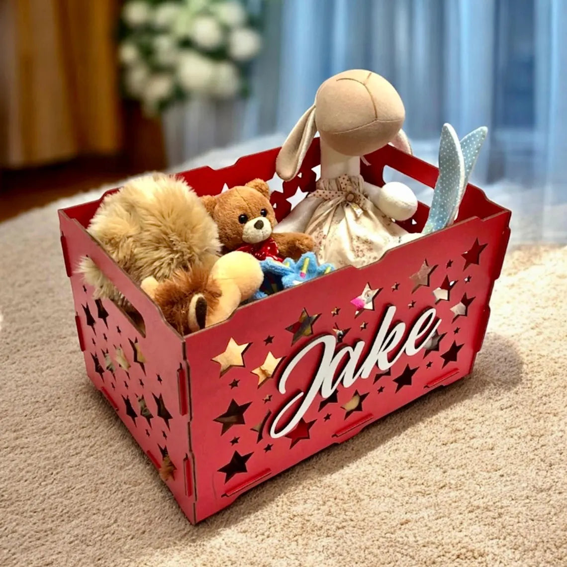 Red toy storage basket with 'Jake' on it, filled with stuffed animals on a carpeted floor.