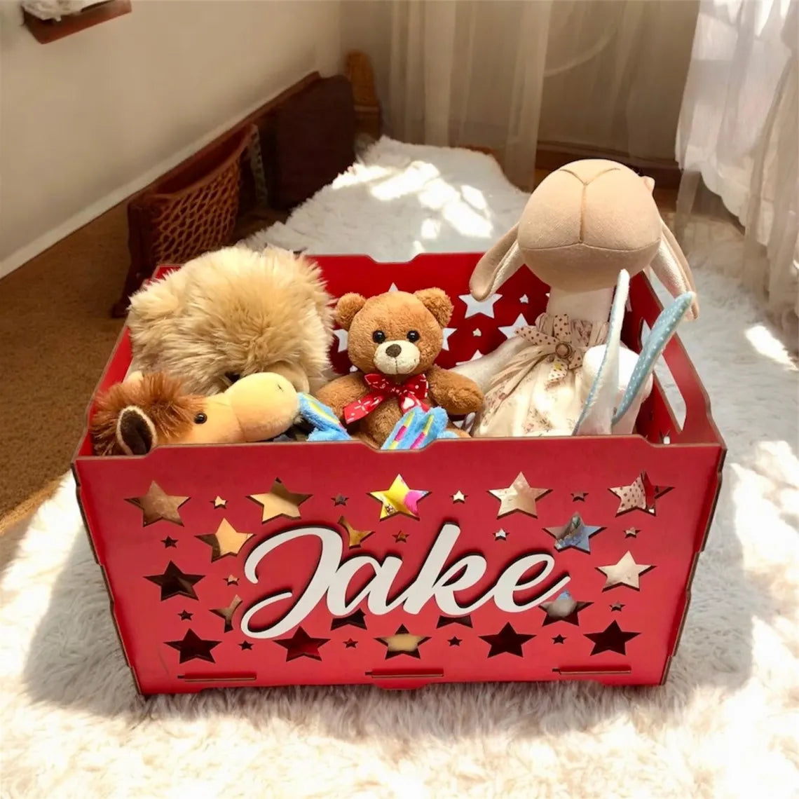 Red toy storage box with 'Jake' on it, filled with stuffed animals, on a carpeted floor.