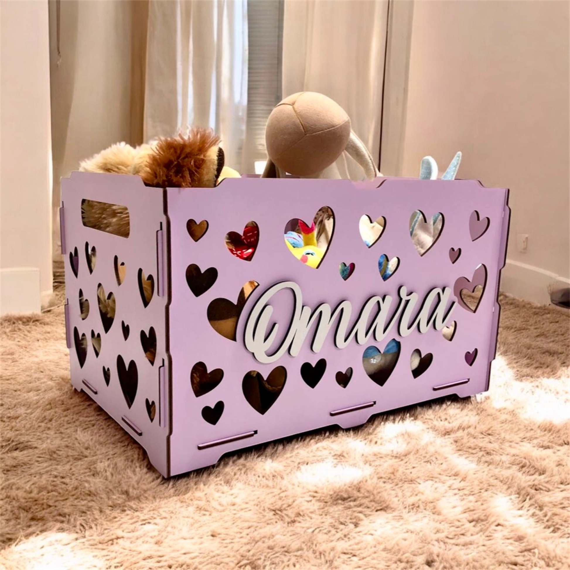 Purple storage crate with heart designs and 'Omara' engraving, containing stuffed animals on a carpeted floor.