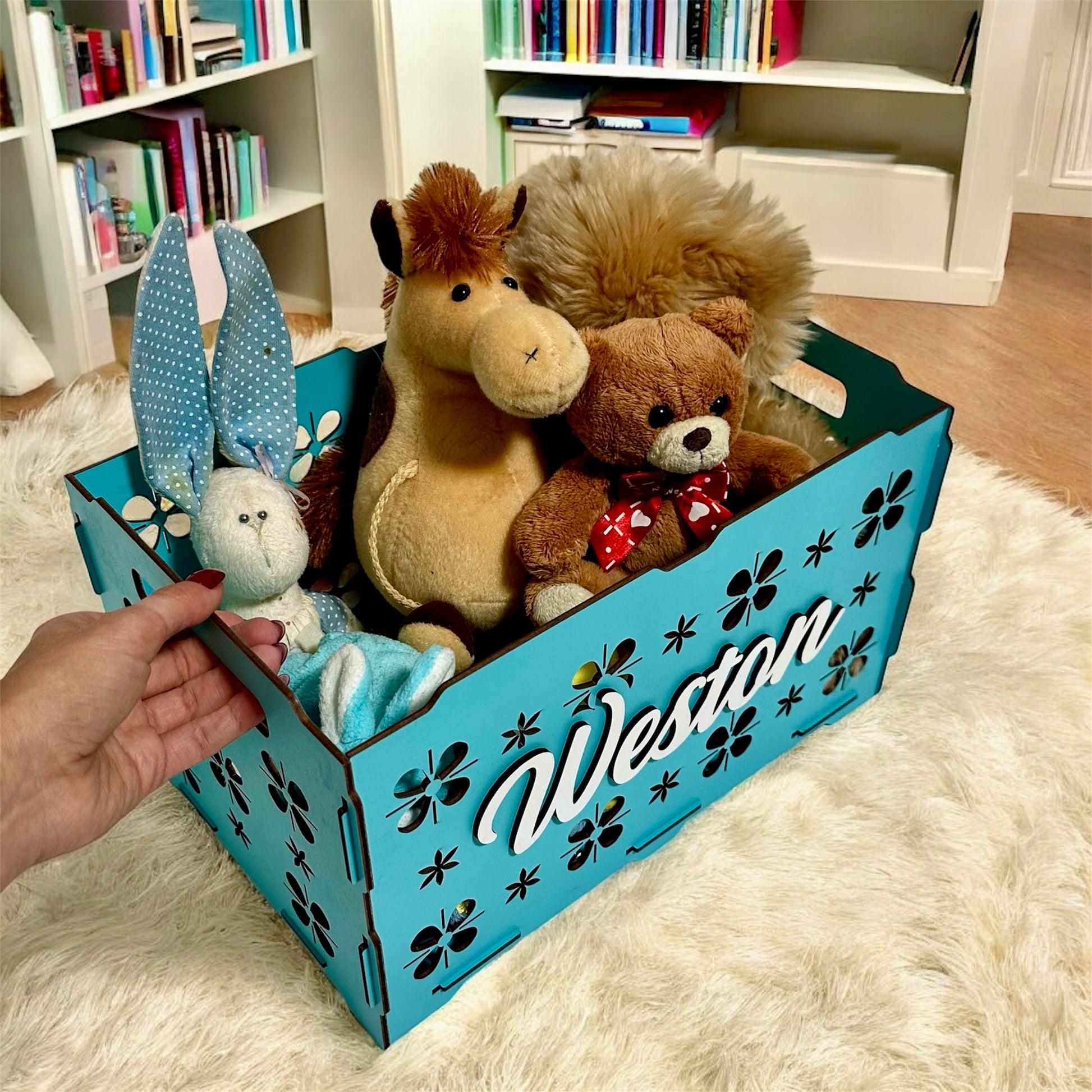 Blue toy storage box with stuffed animals inside, held by a hand on a fluffy rug.