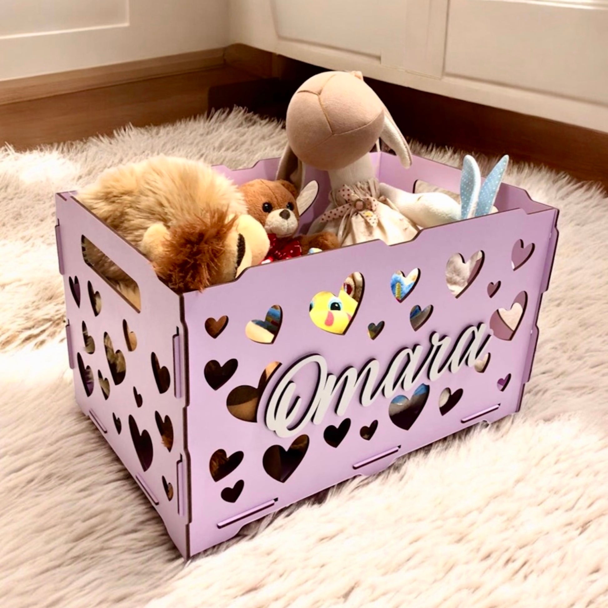 Pink storage crate with heart designs and stuffed toys on a carpeted floor.