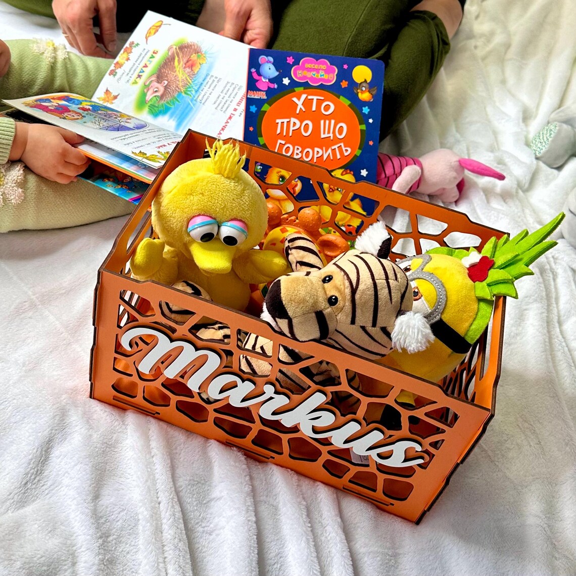 Children's toys and books in a decorative box with 'Markus' on it, on a white blanket.